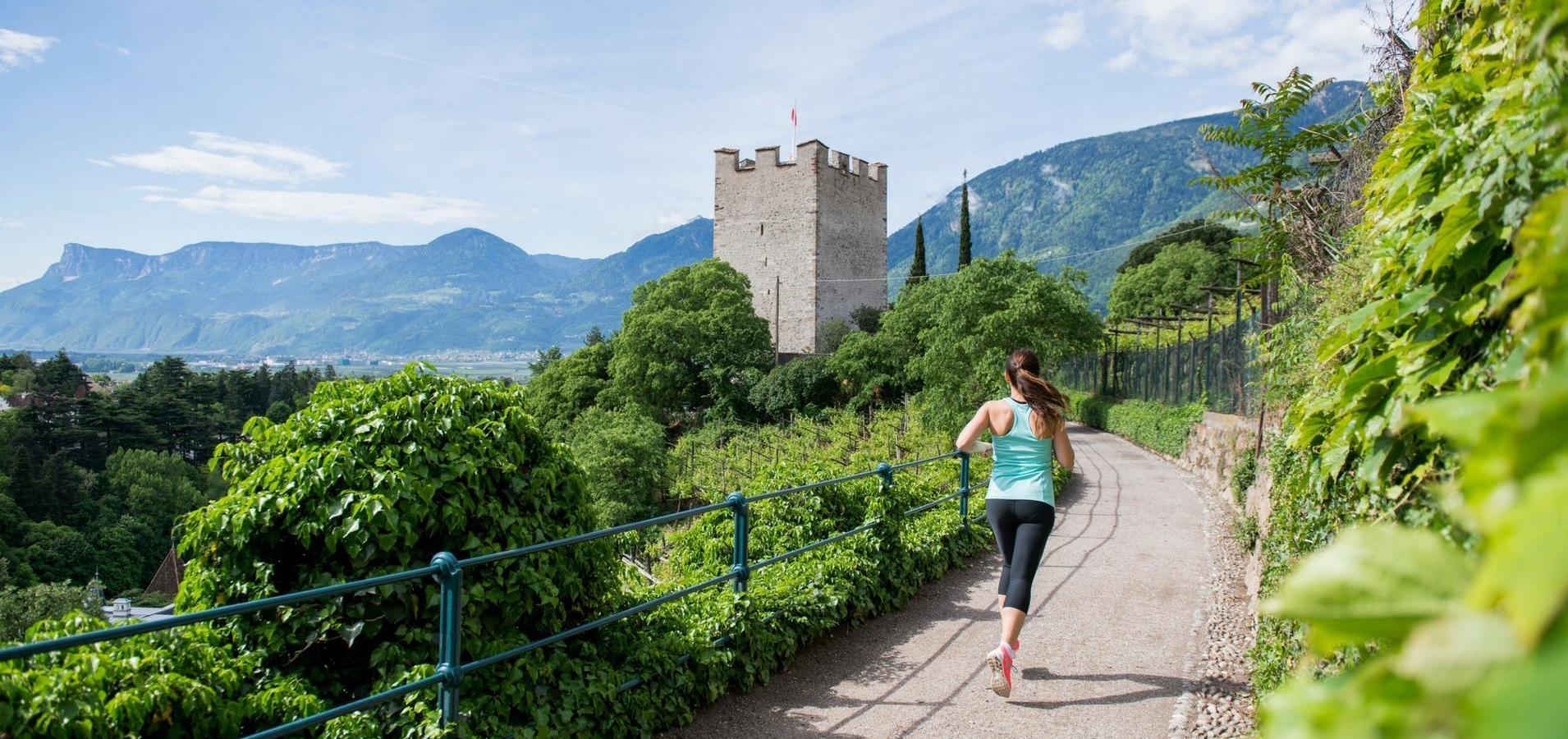 A woman jogging on a scenic path with a castle tower and mountains in the background.